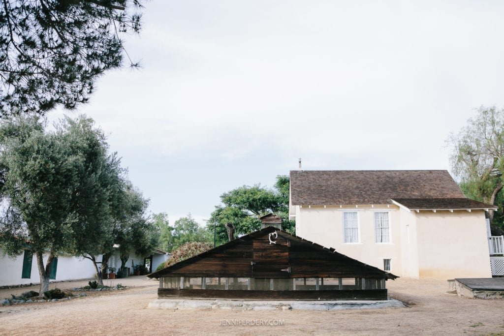 An outdoor scene at Rancho Guajome Adobe featuring an old wooden structure with a slanted roof in the foreground, and a simple, white, single-story building with a brown shingle roof in the background. Trees surround the buildings, and the ground appears dusty under a partly cloudy sky.