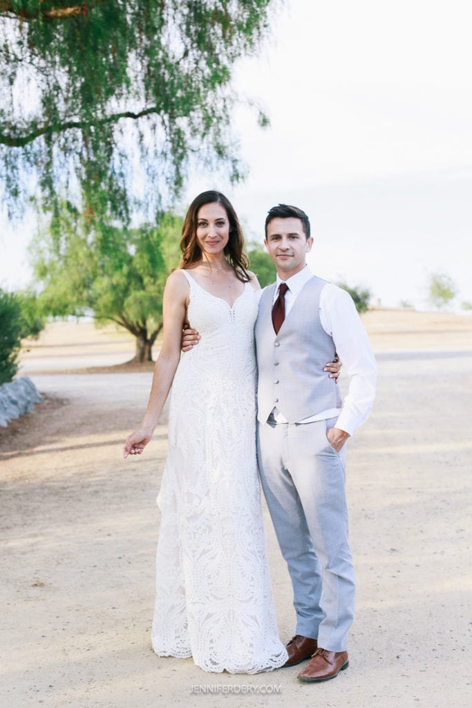A bride and groom pose for a wedding photo outdoors at Rancho Guajome Adobe. The bride is wearing a white lace wedding dress and the groom is dressed in a light gray suit with a white shirt and burgundy tie. They stand on a sandy pathway with trees and greenery in the background.