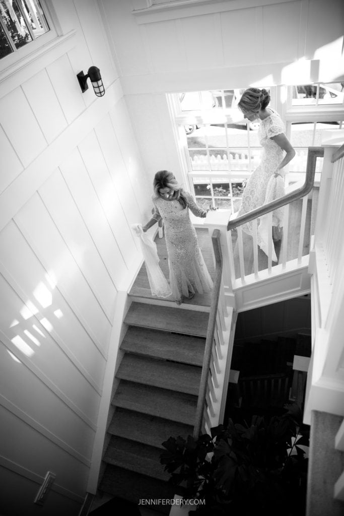 A black-and-white photo of two women in wedding attire walking down a staircase. The woman in the forefront is holding her dress as she descends the stairs, while the other woman looks towards her from behind. Sunlight streams in from a window, casting shadows on the wall.