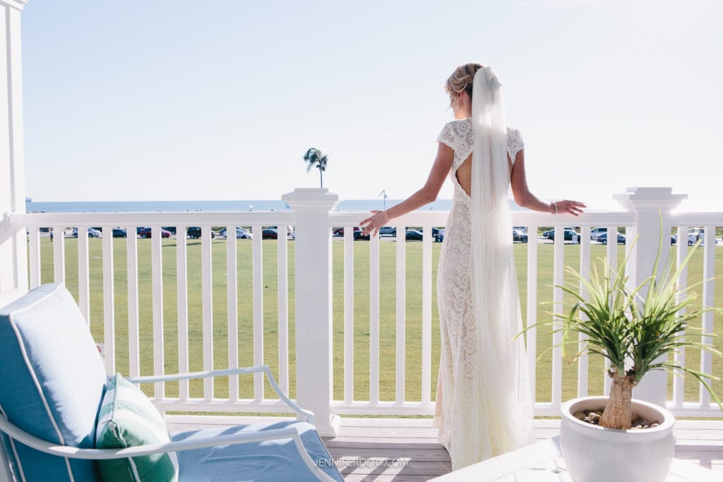 A bride in a white lace gown stands on a balcony with a veil cascading down her back, looking out at a green lawn and distant ocean view. The balcony is adorned with white railings, and patio furniture is placed nearby. A potted plant sits on the ground.