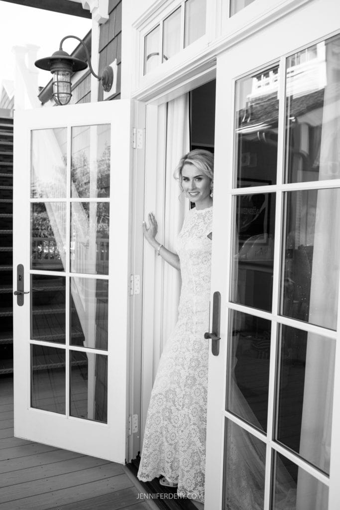 A bride in a lace wedding dress stands in a doorway of a private estate, looking towards the camera with a smile. The door is partially open, and she holds the frame with one hand. The background includes stairs and part of the building. The photo is in black and white.