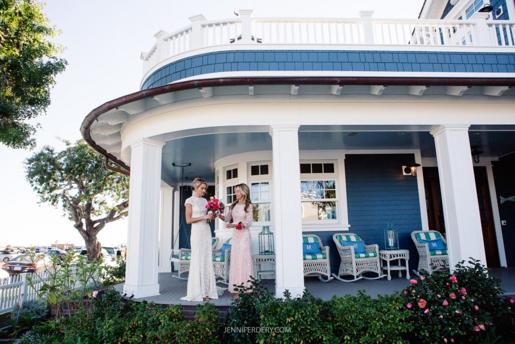 Two women in elegant dresses stand on the porch of a blue and white private home. One holds a bouquet of pink flowers. The house features white columns and a wraparound balcony. Several chairs with blue cushions are arranged on the porch. A tree and greenery surround the scene.