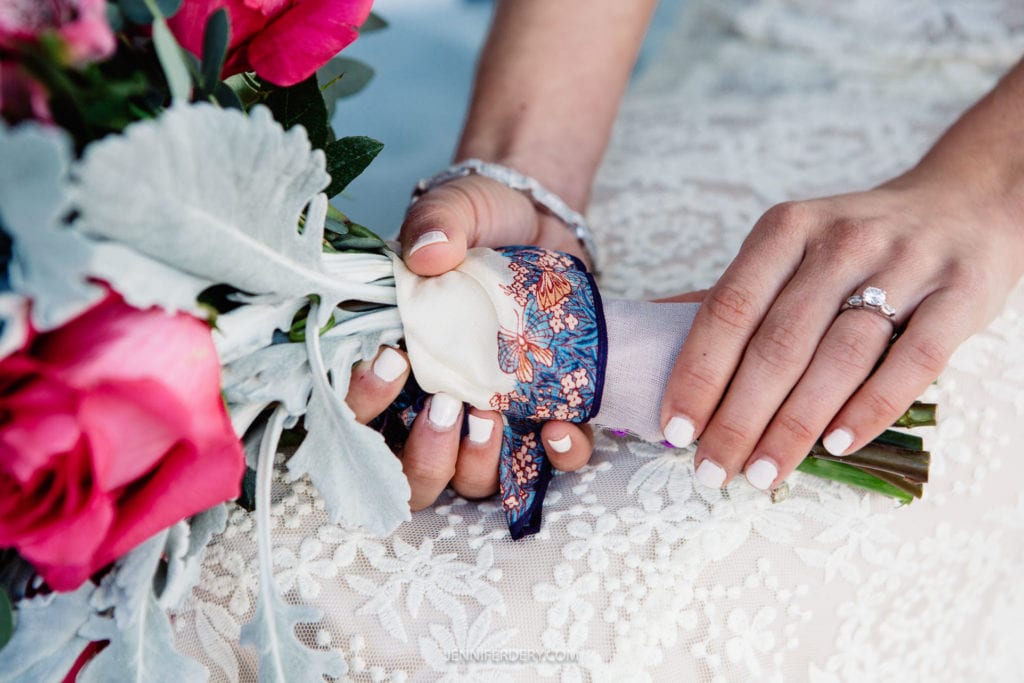 A close-up of a bride's hands holding a bouquet of pink roses and white flowers. Her nails are painted white, and she wears a ring on her left hand. The bouquet is wrapped in a patterned cloth, and she rests it on a lace-covered surface.