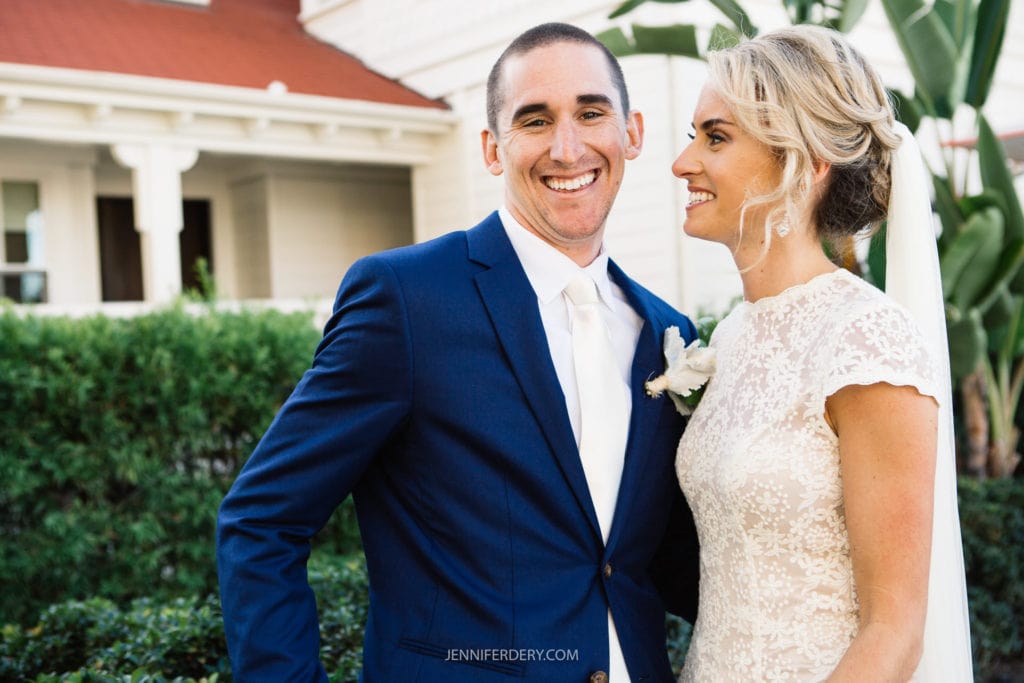A smiling couple dressed in wedding attire stands outside in front of greenery and a building with a red roof. The groom is wearing a blue suit and white tie, while the bride is in a lace dress. The bride looks at the groom while he looks at the camera.
