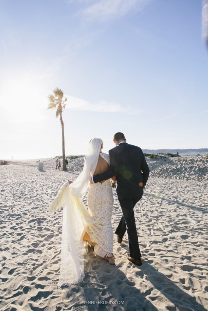 A bride in a white dress and a groom in a dark suit walk arm in arm on a sandy beach with the sun setting behind them. The bride's veil flows in the breeze, and a lone palm tree is visible in the background. The couple is viewed from behind.