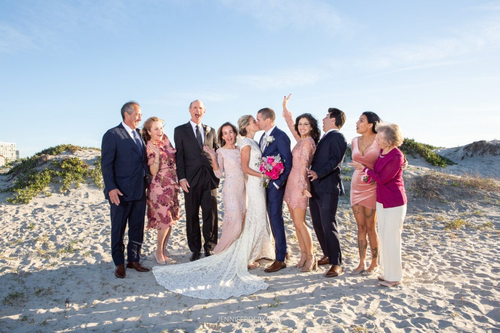 A group of people dressed in formal attire stand on a sandy beach, celebrating a wedding. The bride and groom share a kiss in the center, surrounded by joyful friends and family. Everyone looks happy, with some smiling and raising their hands in excitement.