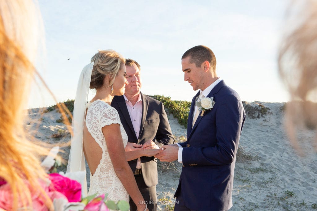 A bride and groom are exchanging rings at a beach wedding. The bride is wearing a white lace dress with an open back, and the groom is in a blue suit with a white rose boutonniere. A officiant stands between them, with sand dunes and a clear sky visible in the background.