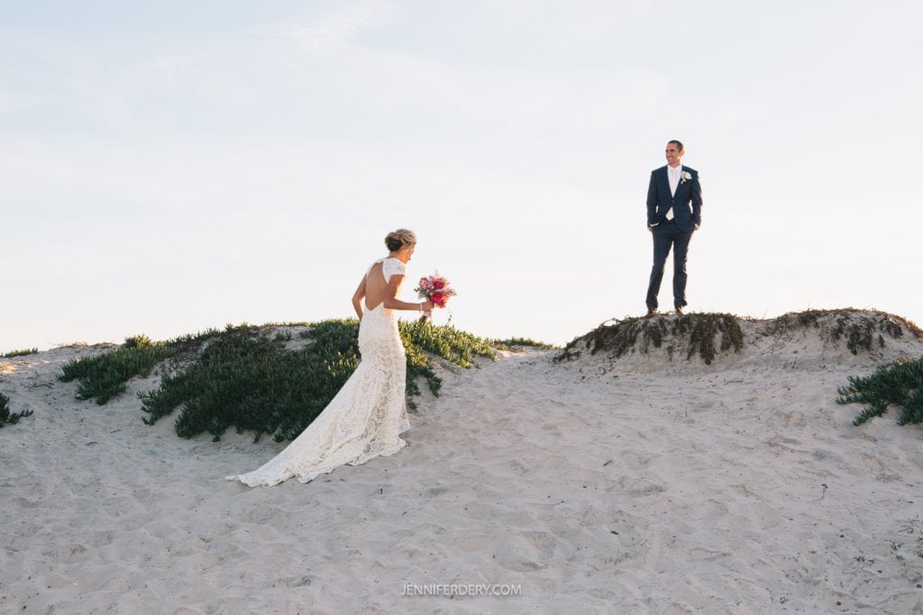 A bride in a lace gown walks up a sandy hill on coronado holding a bouquet of pink flowers. The groom, dressed in a dark suit, stands atop another sandy hill, looking at her. The setting is a beach with clear skies overhead.