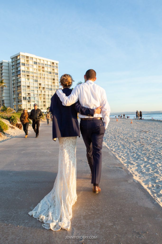 A couple is walking arm-in-arm on a beachside path at sunset. The woman, in a white lace gown and a dark jacket, and the man, in a white shirt and dark pants, are walking away from the camera. A tall building and other people are visible in the background.