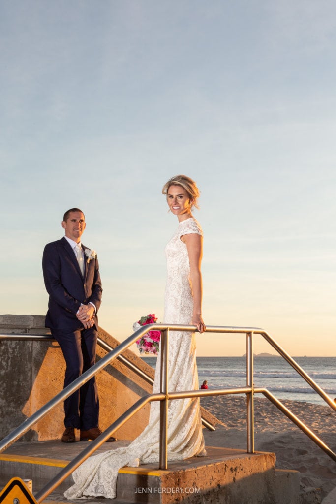 A bride in a white lace gown and groom in a blue suit pose on a beach staircase at sunset. The bride holds a bouquet of pink flowers and looks towards the camera, while the groom stands behind her, gazing into the distance.