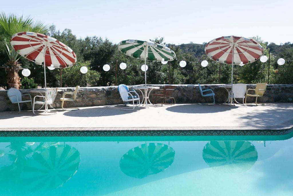 A serene outdoor pool scene, perfect for an intimate elopement, with a row of empty chairs and tables under colorful striped umbrellas. The pool reflects the umbrellas, and string lights add charm to the stone wall backdrop lined with greenery.