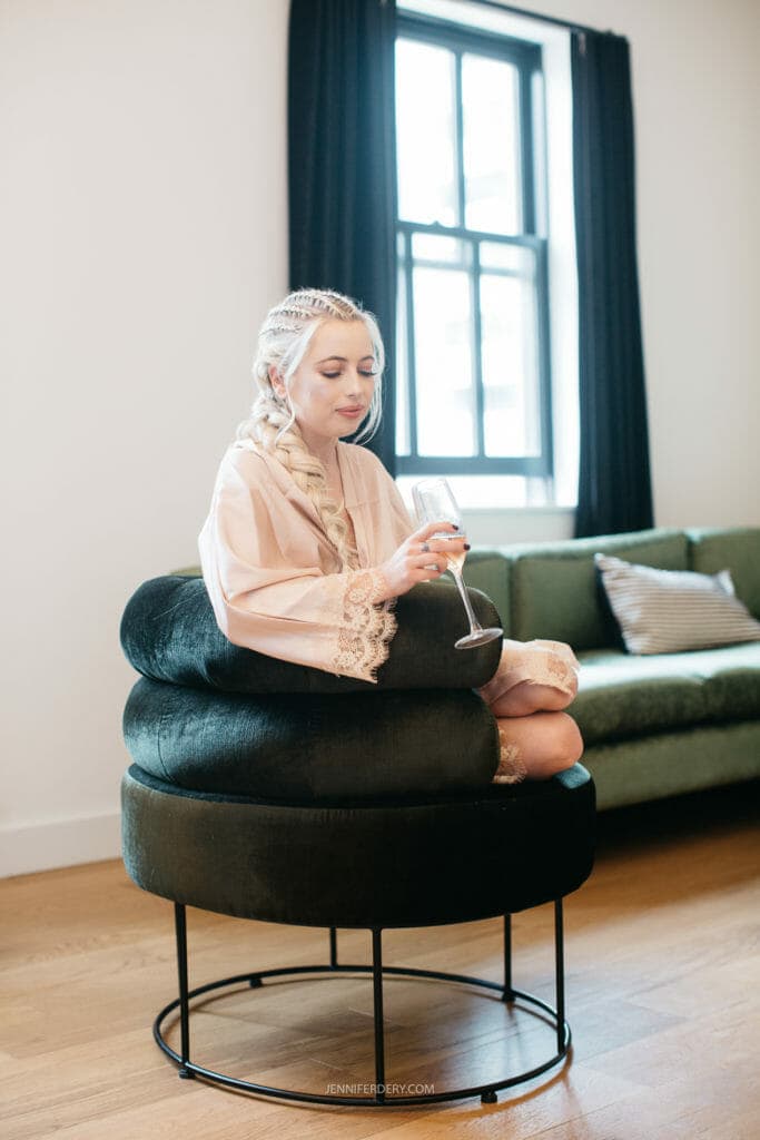 getting ready photo of A bride with long blonde hair in braids, wearing a light pink robe with lace, sits cross-legged on a large black cushioned chair while sipping from a wine glass. She appears relaxed, with a window and green couch in the background.