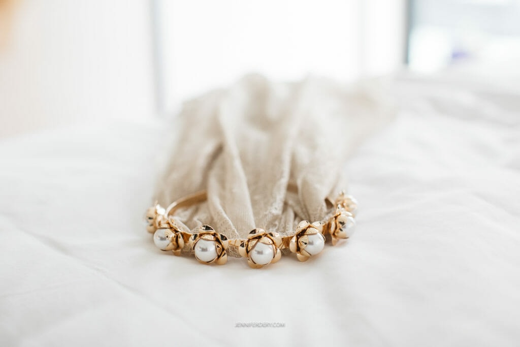 vintage bridal veil: A gold headband adorned with large white pearls and smaller gold beads is placed on a bed with a white bedsheet. The headband is draped with a delicate, sheer, beige fabric. The background is softly blurred, highlighting the headband as the focal point.
