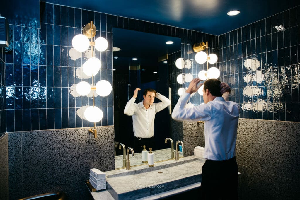 A groom in a white dress shirt is standing in front of a mirror in a stylish, dark-tiled bathroom with bright globe lights. He appears to be fixing his hair. The bathroom countertop has a soap dispenser and neatly stacked towels.
