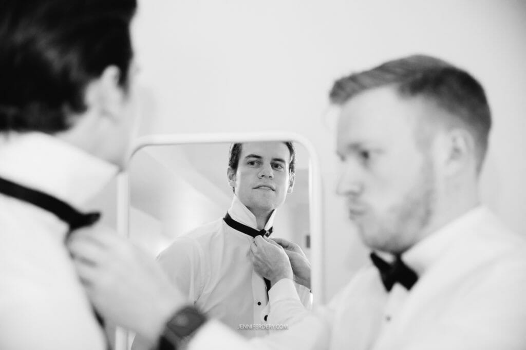 Two men wearing formal attire are adjusting their bow ties in front of a mirror. The man on the left, with dark hair, is intently tying his bow tie, while the man on the right, with light hair, is focused on perfecting his own. Both are dressed in white dress shirts.