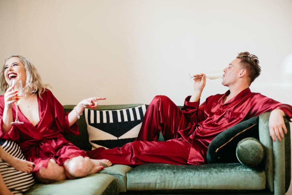 Two people are sitting on a green couch wearing matching red satin pajamas. On the left, a person laughs heartily while pointing and holding a champagne glass. On the right, another person lounges, sipping champagne. The background features a white wall.