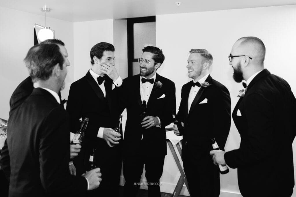 A group of six men in tuxedos stand in a room, laughing and chatting. One man covers his mouth with his hand while holding a beer. Another man gestures with his hand, also holding a beer. They appear to be in a celebratory or pre-event setting.