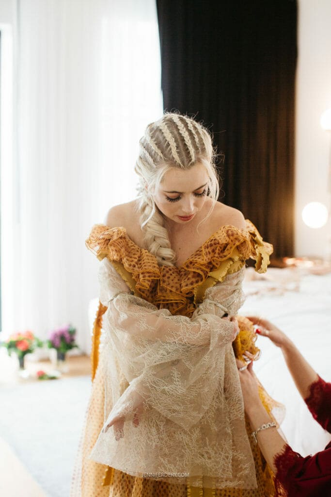 A bride with light blonde daenerys khaleesi braided hair is adjusting her intricate, vintage-inspired dress with lace and ruffles. She appears to be in a well-lit room with a bed, flowers, and a black curtain in the background.