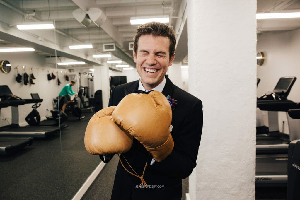A groom wearing a black suit and a bow tie stands in a gym with a wide smile, holding up oversized boxing gloves in a playful manner. Exercise equipment, such as treadmills, is visible in the background.