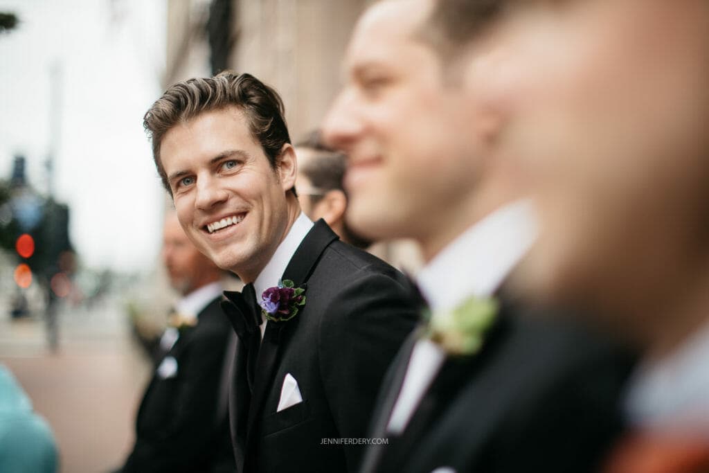 A groom smiles while sitting with his groomsmen. He’s wearing a black tuxedo with a purple boutonniere. The background is slightly blurred, showing an outdoor urban setting with soft lighting. Other groomsmen in similar attire are visible but out of focus.