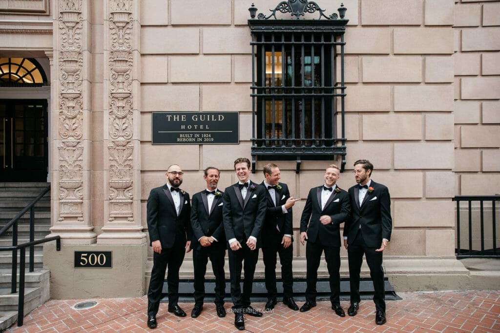 A group of six groomsmen dressed in formal black suits and bow ties stand outside The Guild Hotel. They are smiling and laughing together. Behind them is a stone building with ornate details and a sign reading "The Guild Hotel". The address number 500 is visible.