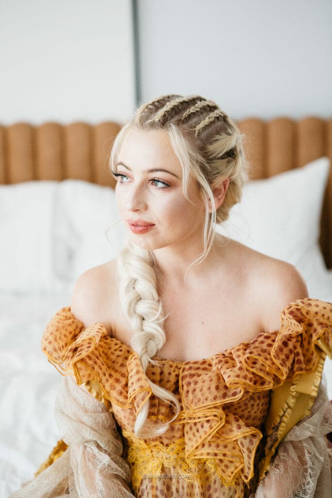 A bride with long, blonde hair styled in a braid and cornrows sits on a bed with white pillows. She is wearing an off-shoulder, ruffled top with a yellow and orange polka-dot pattern. She looks slightly to her left with a calm expression.