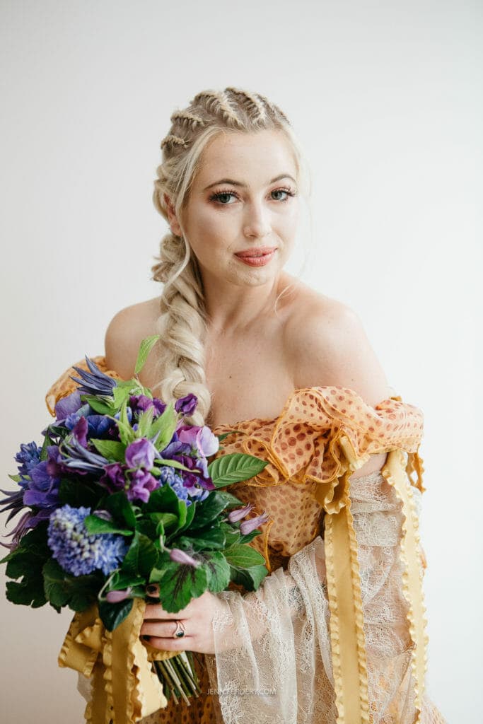 A bride with long, braided blonde hair wearing a yellow and orange off-the-shoulder dress holds a bouquet of vibrant purple flowers. She gazes softly at the camera, standing against a plain white background.