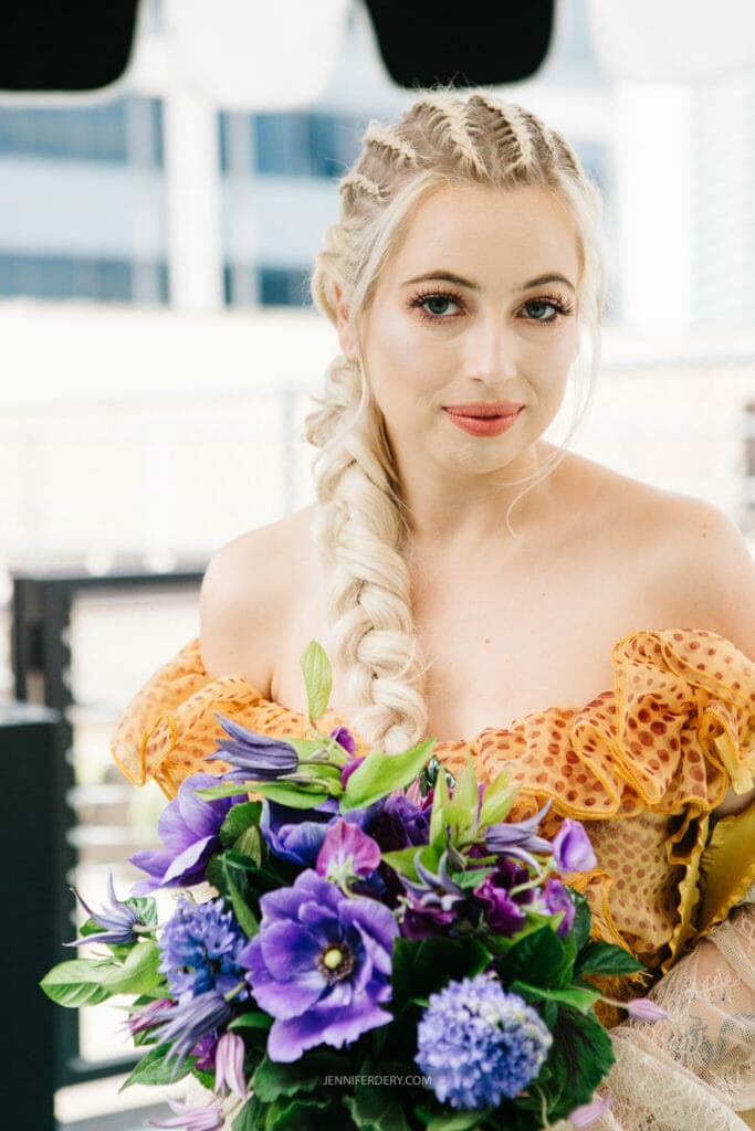A bride with blonde braided hair and light makeup is wearing an off-the-shoulder, colorful dress. She is holding a bouquet of vibrant purple and blue flowers with green leaves. The background is an outdoor setting with blurred urban buildings.