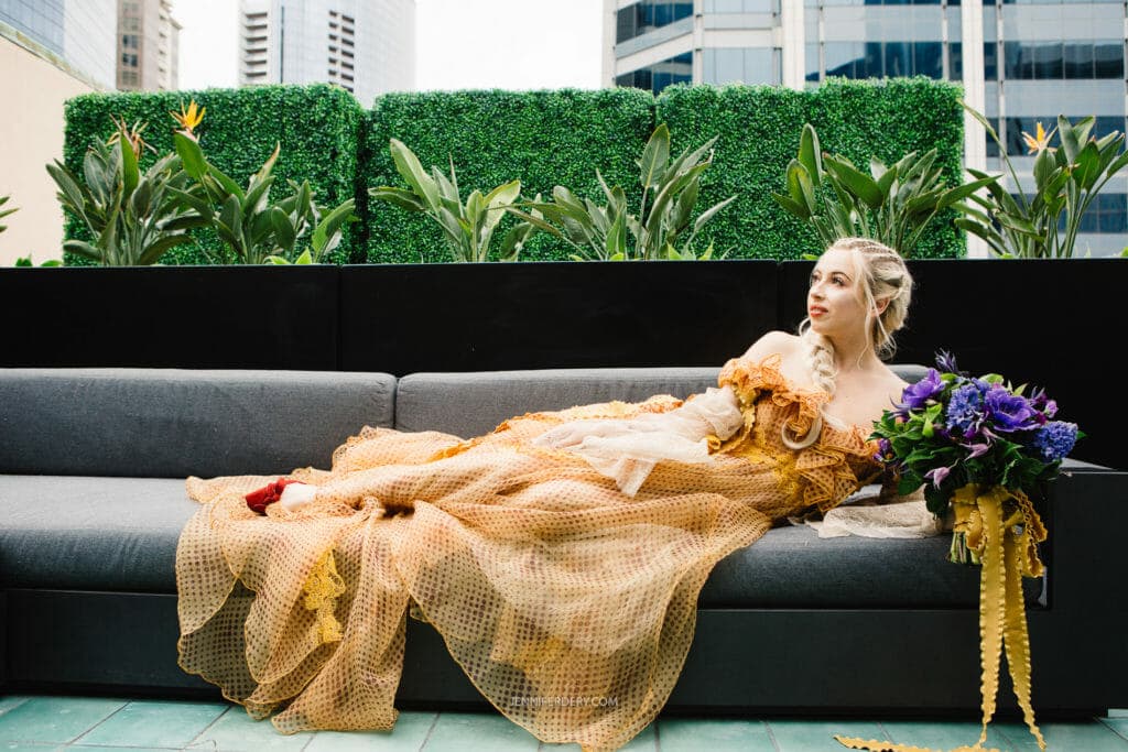 A bride wearing a flowing gold-colored dress with mesh-like details reclines on a gray outdoor sofa, holding a large bouquet of vivid purple flowers. Behind her is a black divider adorned with lush green foliage, and tall buildings are visible in the background.