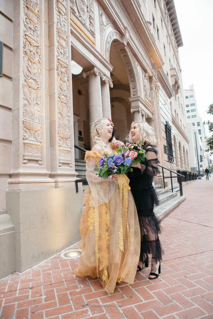 Two women stand outside a decorative building, laughing and holding a colorful bouquet of flowers. One woman is dressed in a gold gown with detailed patterns, while the other wears a black dress with sheer overlays. The scene is lively and joyful.
