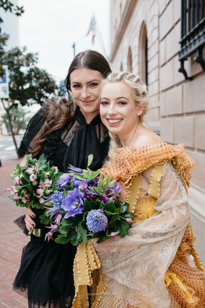 Two women in elegant dresses stand outside a building on a city street. One wears a black dress while the other is in a yellow dress with lace. Both smile and hold vibrant bouquets of flowers, including purple lilies and blue hydrangeas. Trees and a flag are in the background.