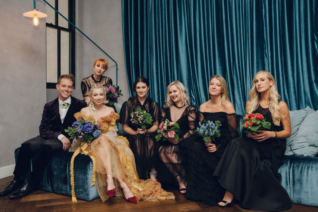 A group of seven people, five women and two men, are sitting on a blue velvet couch holding bouquets of flowers. They are dressed in formal attire and smiling at the camera. The background features dark teal curtains and modern lighting.