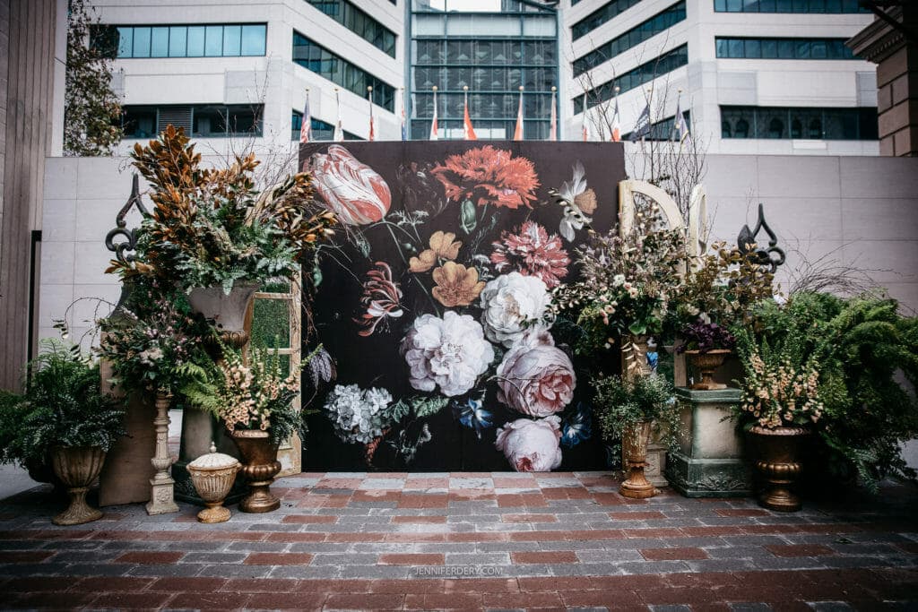 An outdoor display features a large floral mural with various flowers, including roses, tulips, and carnations. Surrounding the mural are numerous potted plants and decorative planters on a brick-paved area, with tall buildings and a flag in the background.