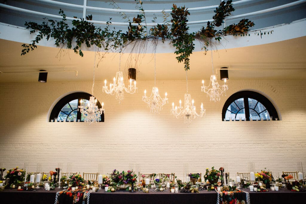 A long dining table adorned with vibrant floral arrangements and candles sits beneath elegant chandeliers. Above, green foliage and other greenery decorate the white ceiling. Two circular windows in the background let in natural light.