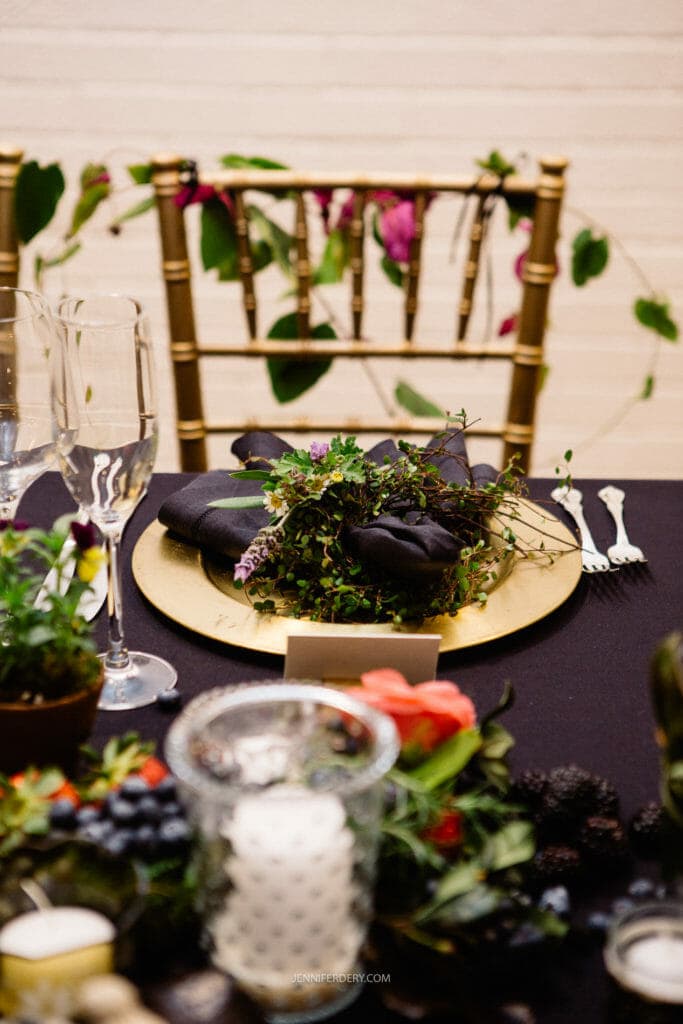 A close-up of an elegantly set table with a gold chair, black linens, and a gold charger plate topped with black napkins and greenery. The centerpiece includes candles and an arrangement of orange and dark purple flowers. Crystal glassware is also present.