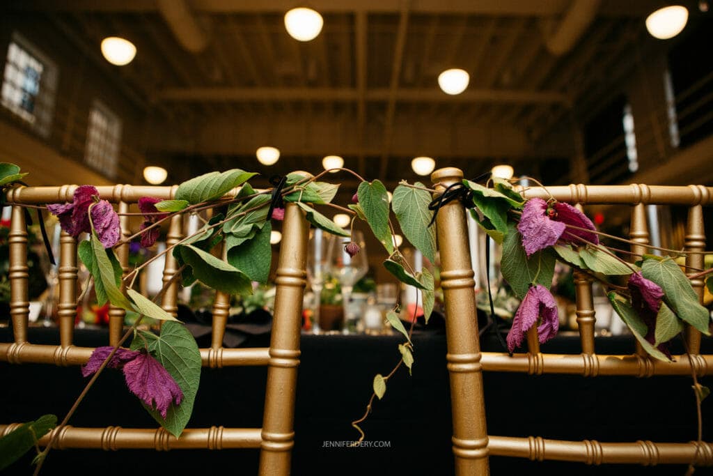 Golden chairs adorned with a garland of green leaves and purple flowers are situated in a warmly lit indoor space, possibly a venue for an event like a wedding or banquet. The background shows blurred tables and round ceiling lights.