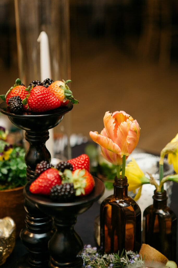 A decorative arrangement featuring strawberries and blackberries on black candleholders and a vase with an orange tulip. Brown glass bottles hold additional flowers in the background, creating a colorful display.