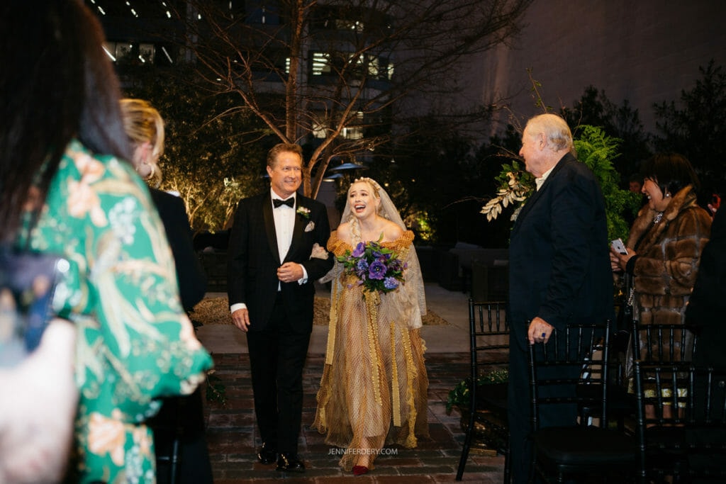 A bride, wearing a long gold dress, walks down an outdoor aisle with her father, who is in a black tuxedo. She holds a vibrant bouquet and appears joyful. Guests, some in formal attire, watch them under soft evening lighting. Trees and a building are in the background.