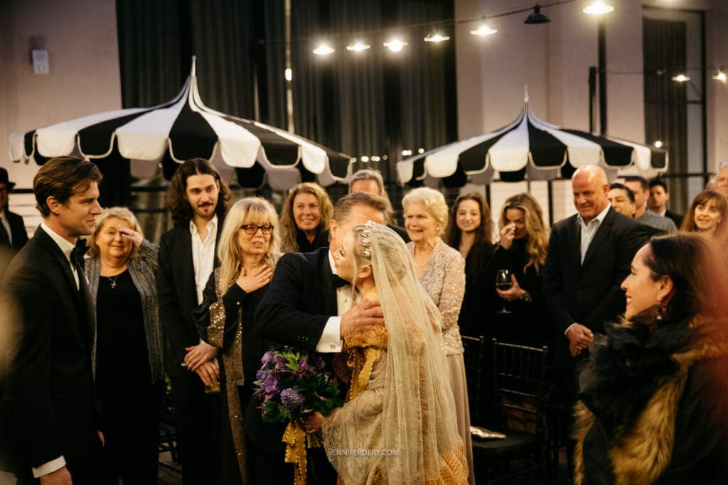A bride in a white gown and veil hugs an older man in a suit while holding a bouquet of purple flowers. They're surrounded by smiling guests in formal attire under black and white canopy tents. The room is warmly lit, creating a joyful atmosphere.