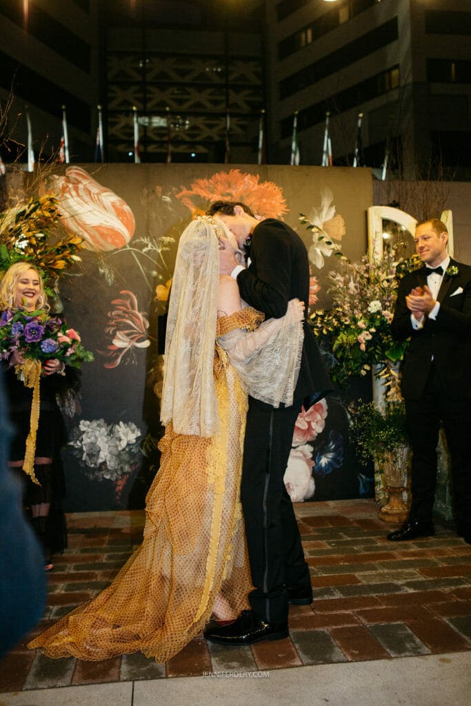 A newlywed couple shares a passionate kiss at their outdoor wedding ceremony. The bride wears a unique, golden-hued lace dress with a matching veil, while the groom is in a black suit. They stand in front of a floral backdrop, with bridesmaids and groomsmen looking on joyfully.