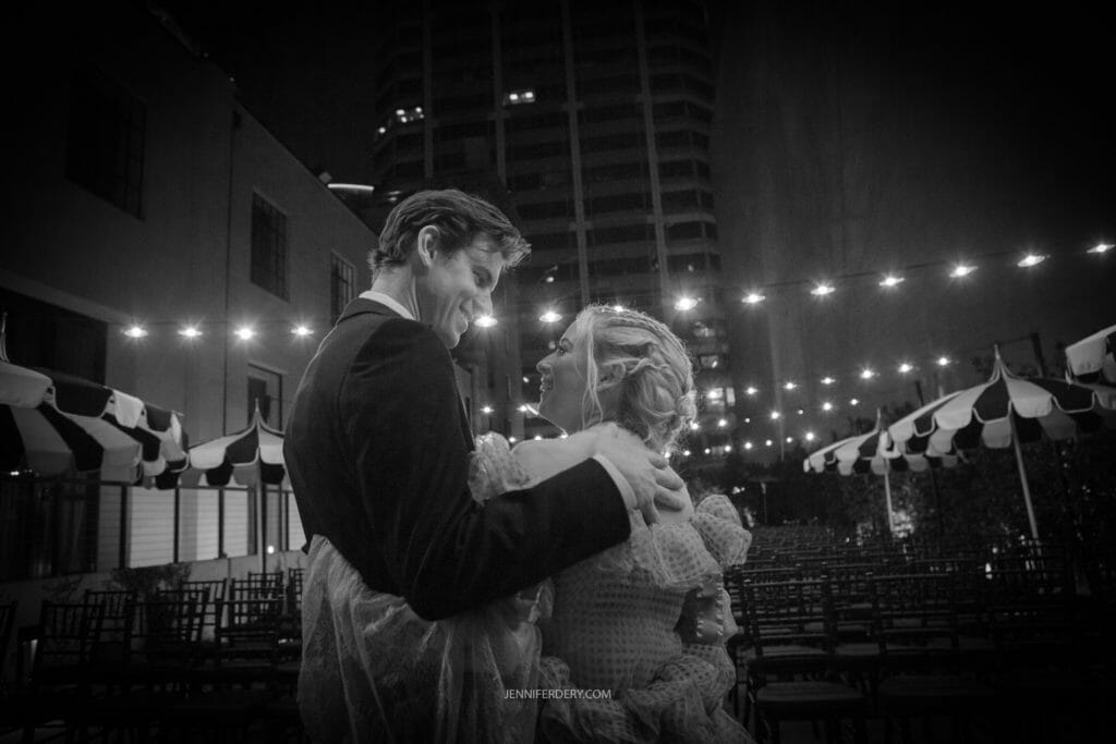 A couple dressed in wedding attire shares a joyful moment, gazing into each other's eyes while dancing at an outdoor night venue. String lights and striped canopies enhance the romantic atmosphere. They appear to be celebrating their wedding.
