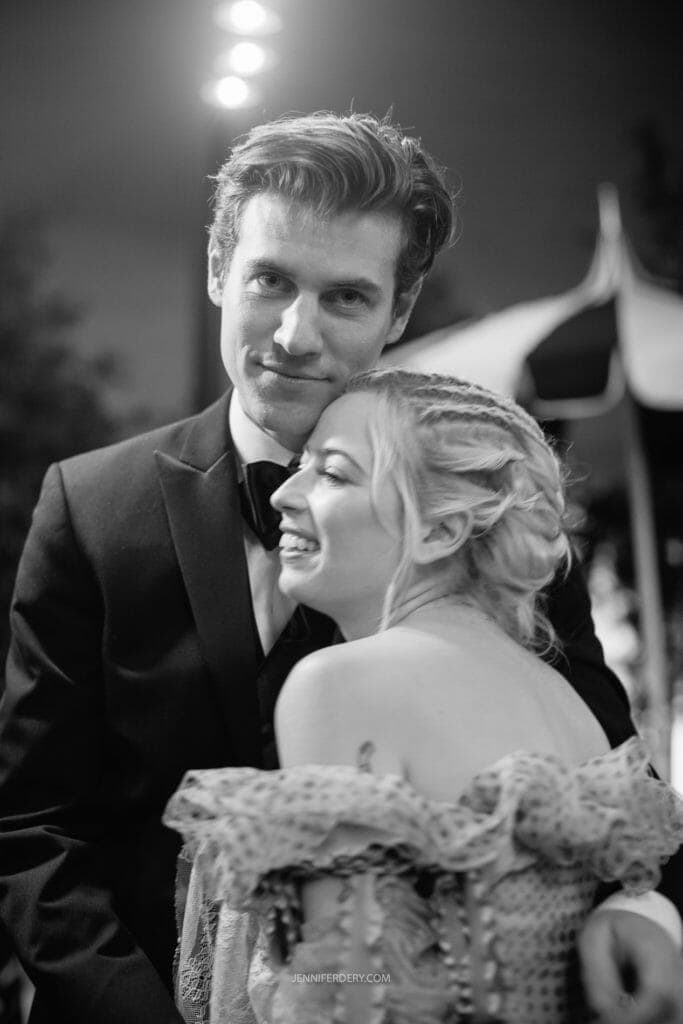 A black and white photo of a smiling couple embracing at an event. The man, wearing a tuxedo, gazes at the camera. The woman, with braided hair and an intricate off-shoulder dress, leans against him, looking to her right. The background is softly blurred.