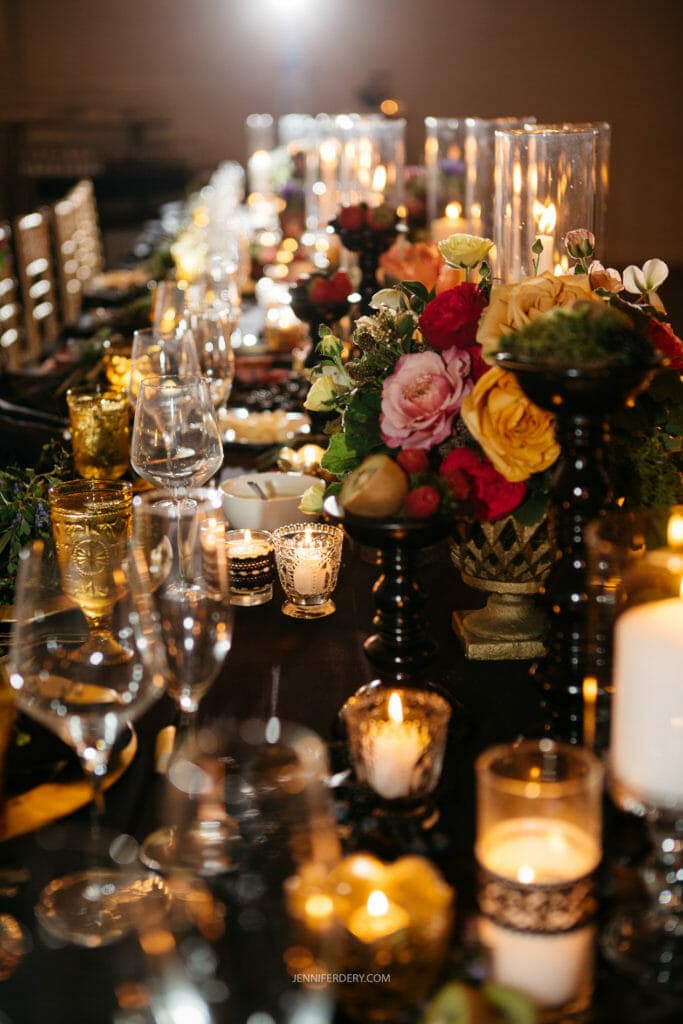 A long dining table is elegantly set with lit candles, vibrant flower arrangements, and neatly arranged glassware. The table's decor includes gold accents and a variety of floral centerpieces, creating a warm and inviting atmosphere suitable for a formal event or celebration.