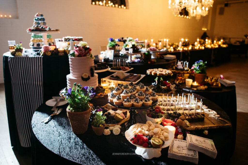 A beautifully decorated dessert table at a wedding reception. Various sweets, including cupcakes, pastries, and cake pops, are elegantly arranged. A multi-tiered wedding cake and a potted plant in the foreground add charm. The background features soft candlelight.