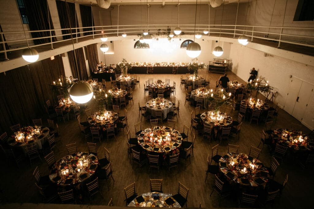 A large, elegantly decorated ballroom at the guild hotel in san digo, with numerous round tables set for dining. Warm lighting illuminates the space, creating a cozy ambiance. Tables are adorned with candles and floral centerpieces. Overhead lights hang from the high ceiling, adding to the sophisticated atmosphere.