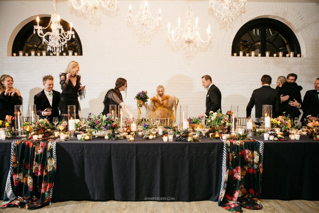 A wedding reception in an elegantly decorated room. The bridal party stands behind a long table adorned with flowers and candles. The bride, in focus, holds her bouquet while interacting with guests. Chandeliers and arched windows enhance the ambiance.
