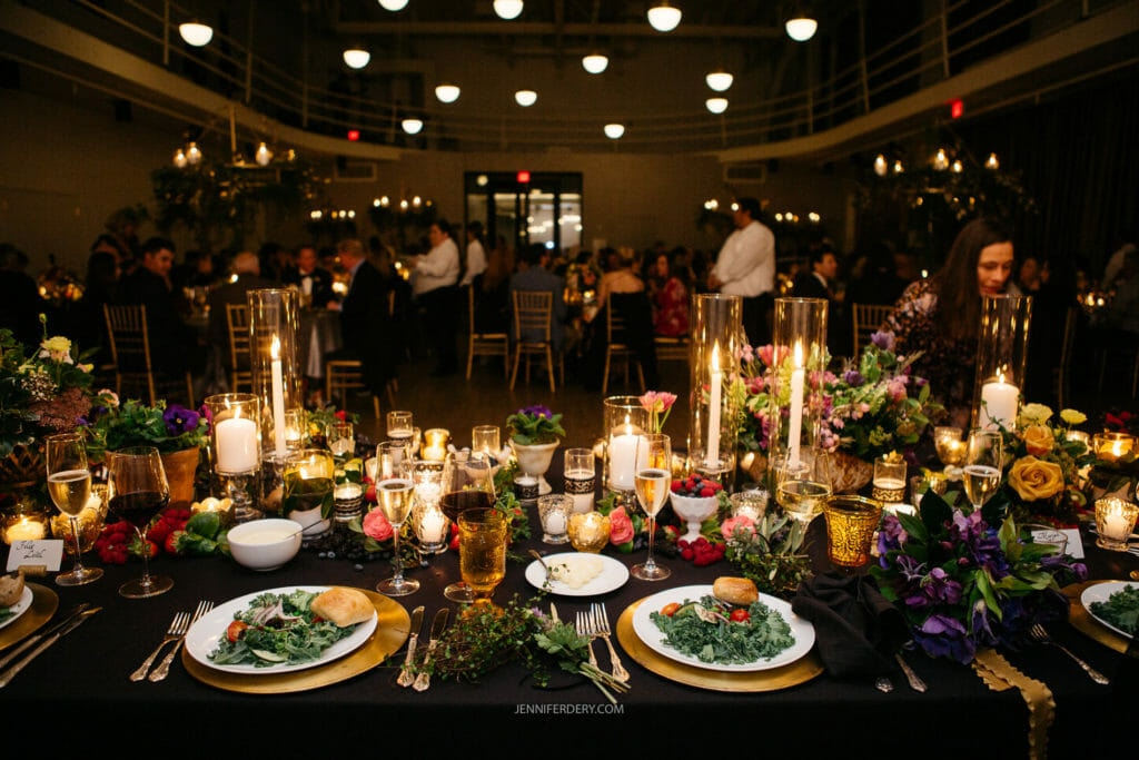 A beautifully decorated dining table set for an elegant dinner event. The table features lit candles, various floral arrangements, glasses of wine, and plated salads. Guests are seated in the background, engaged in conversation under dim, warm lighting.