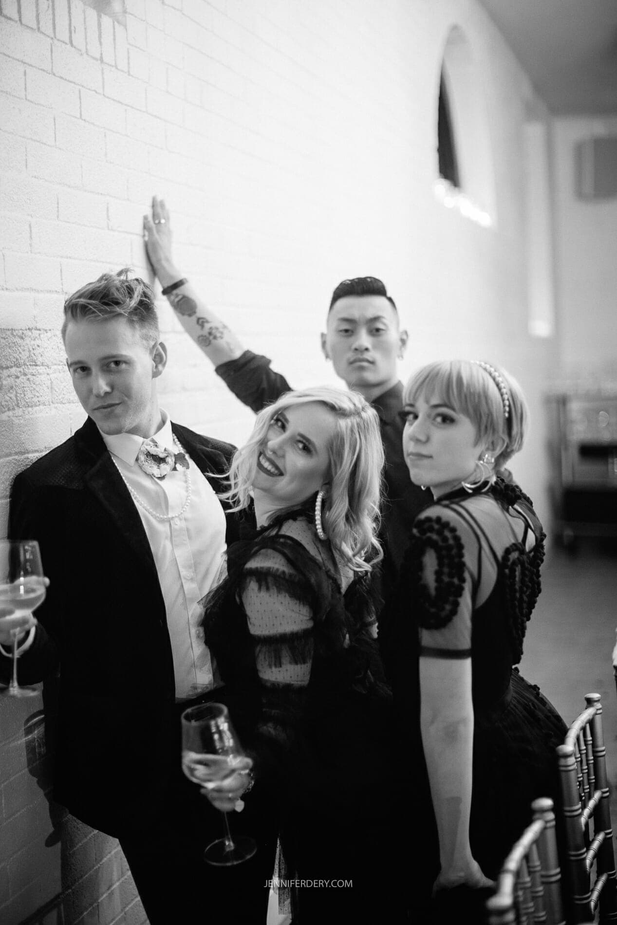 A black-and-white photo shows two men and two women dressed in formal attire, posing and smiling at the camera. They each hold a glass of wine. The setting appears to be an indoor event with white brick walls and large windows.