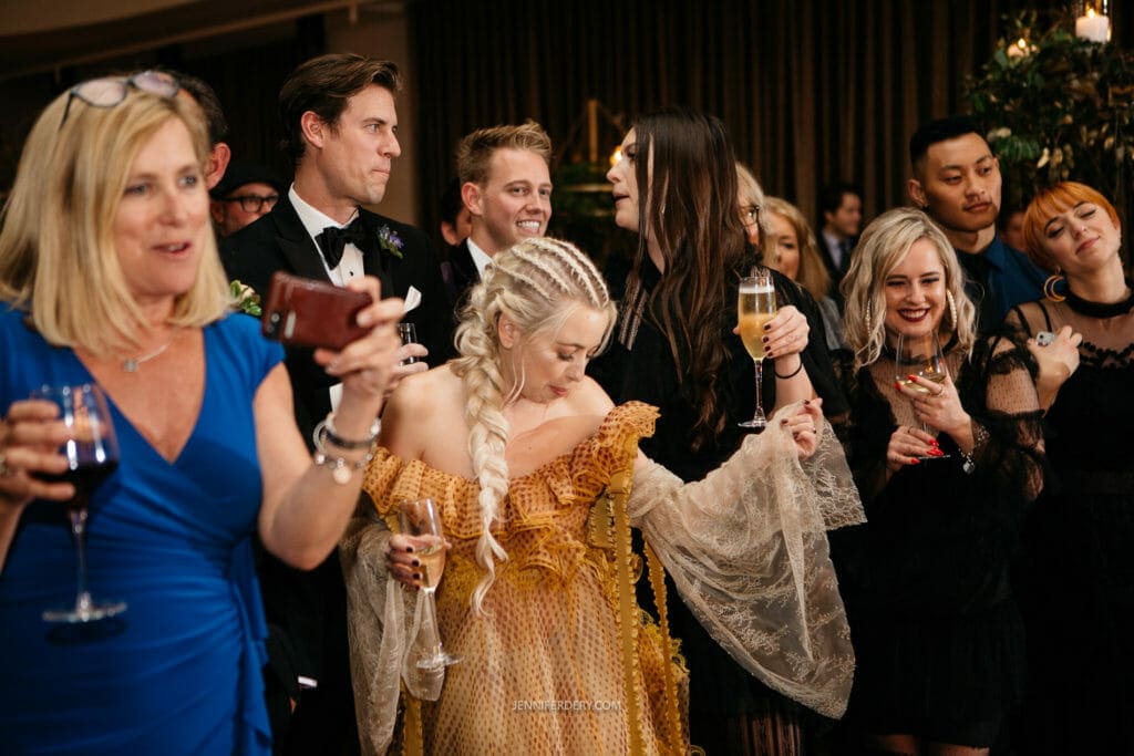 A lively group of people dressed in formal attire are seen at an event. One woman in the foreground wearing a blue dress is holding a wine glass and looking at her phone. Another woman in a golden, flowy dress is in the center, surrounded by smiling and talking individuals.