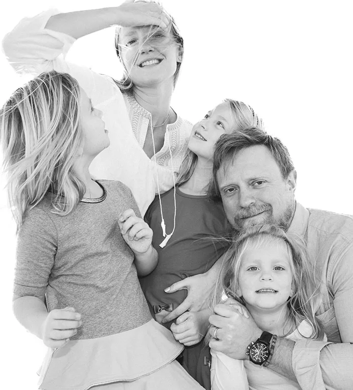A black and white photo of a family. A woman stands at the back, brushing her hair aside, smiling. A man sits in front holding a young girl while another girl stands to the left. Two young girls stand close, looking at the woman. Everyone appears happy.